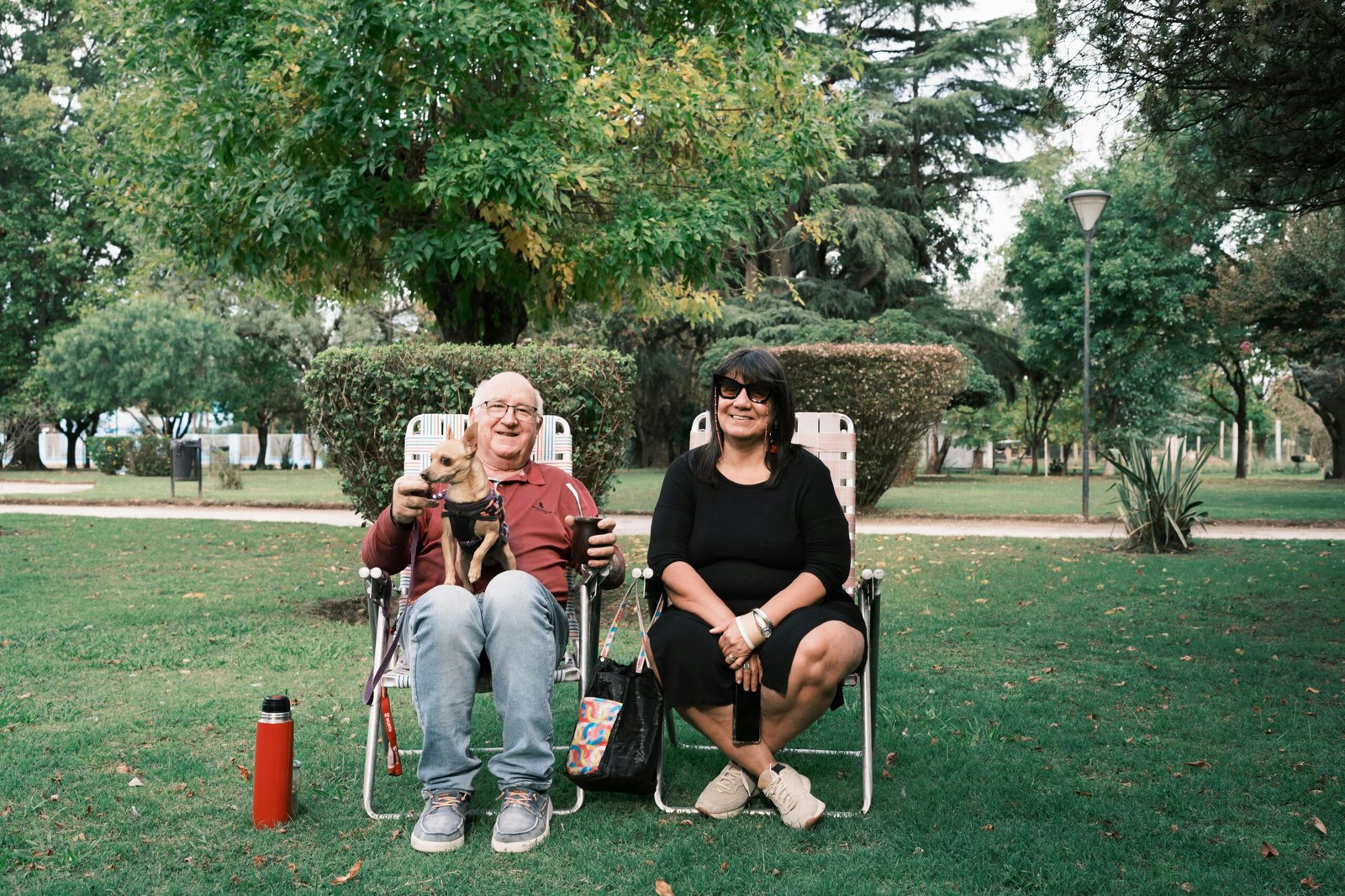Two adults enjoying a sunny day in a park, seated on chairs with their dog, surrounded by greenery.